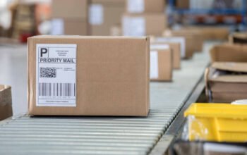 warehousing A brown cardboard box sits on a conveyor belt in a distribution warehouse. It has a priority mail label on it as well as a bar code and QR code.